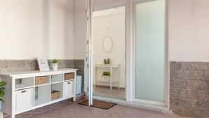Un hall d’entrée propre et moderne, avec une console blanche sur laquelle sont posés des paniers et un panneau d’accueil. Une porte-fenêtre coulissante donne accès à un balcon agrémenté d’une petite table et de plantes en pot.