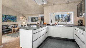 Et moderne køkken med en central ø, hvide skabe og et stort vindue med udsigt til træer.
