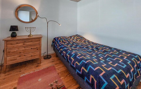 A bedroom with a wooden dresser, a round mirror, a floor lamp, a patterned bedspread, and a red rug.