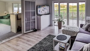Un salon moderne avec une grande télévision, un canapé vert, une petite table et une porte-fenêtre coulissante donnant sur un balcon avec vue sur un champ.