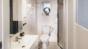 Une petite salle de bain avec un lavabo, des toilettes et une douche, le tout de couleur blanche. Une petite fenêtre située au-dessus des toilettes est décorée d’une guirlande.