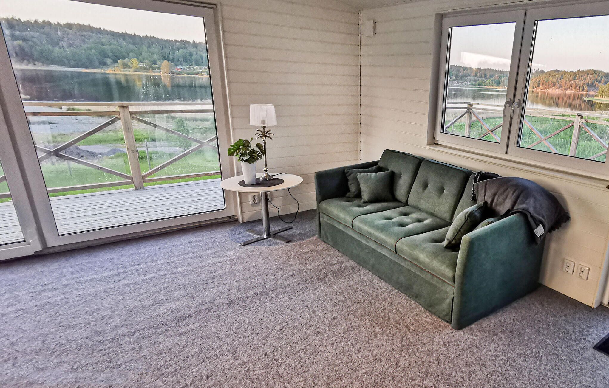 A room with a green sofa, a small table with a lamp, and a view of a lake through large windows.