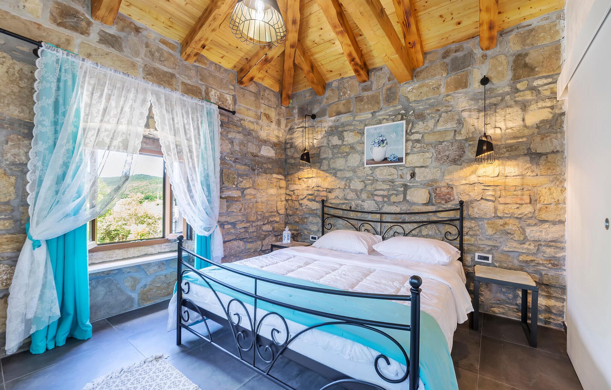 A bedroom with a stone wall, a bed with a metal frame, a nightstand, a window with curtains, and a hanging light fixture.