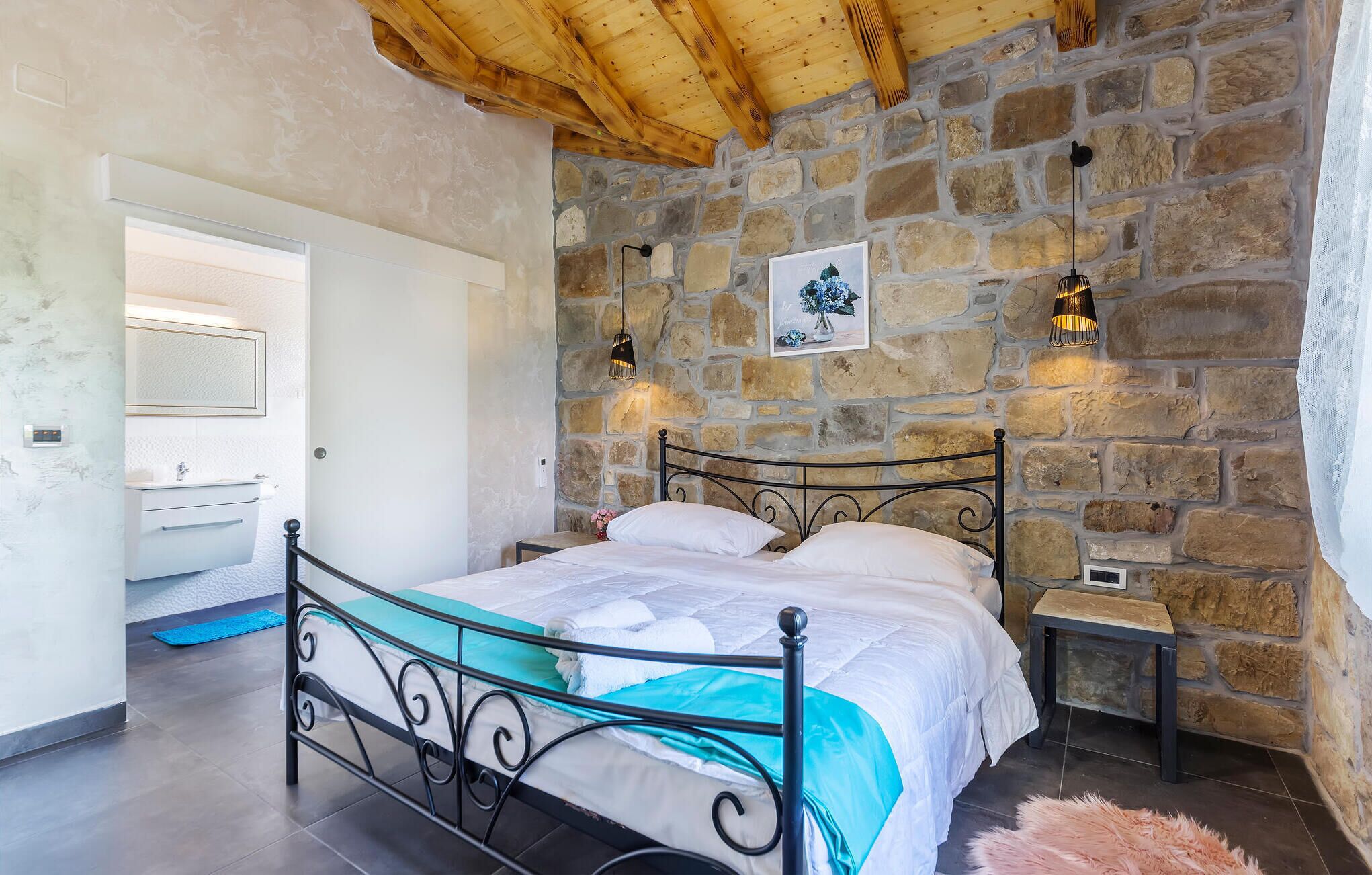 A bedroom with a stone wall, a bed with a metal frame, a bedside table, and a nightstand.