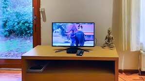 A television on a wooden stand with a remote control, a small statue, and a window with a view of greenery.