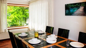 A dining area with a table set for four, a view of greenery through a window, and a painting of a wooden bridge over water on the wall.