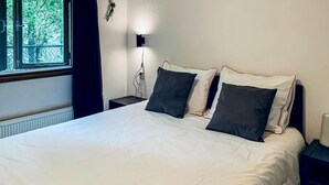 A neatly made bed with white bedding and dark pillows, a bedside lamp, and a window with curtains.