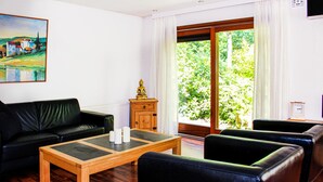 A living room with a black leather sofa, a wooden coffee table, and a wooden cabinet with a Buddha statue. There is a painting on the wall and a window with a view of greenery.