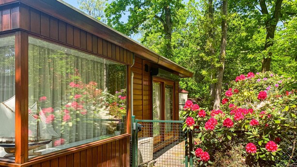 A wooden cabin with large windows, a covered porch, and a garden with blooming flowers.