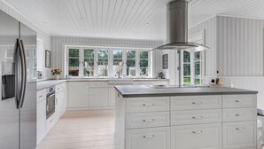 A spacious kitchen with white cabinetry, a large island with drawers, a stainless steel refrigerator, and a view through large windows.