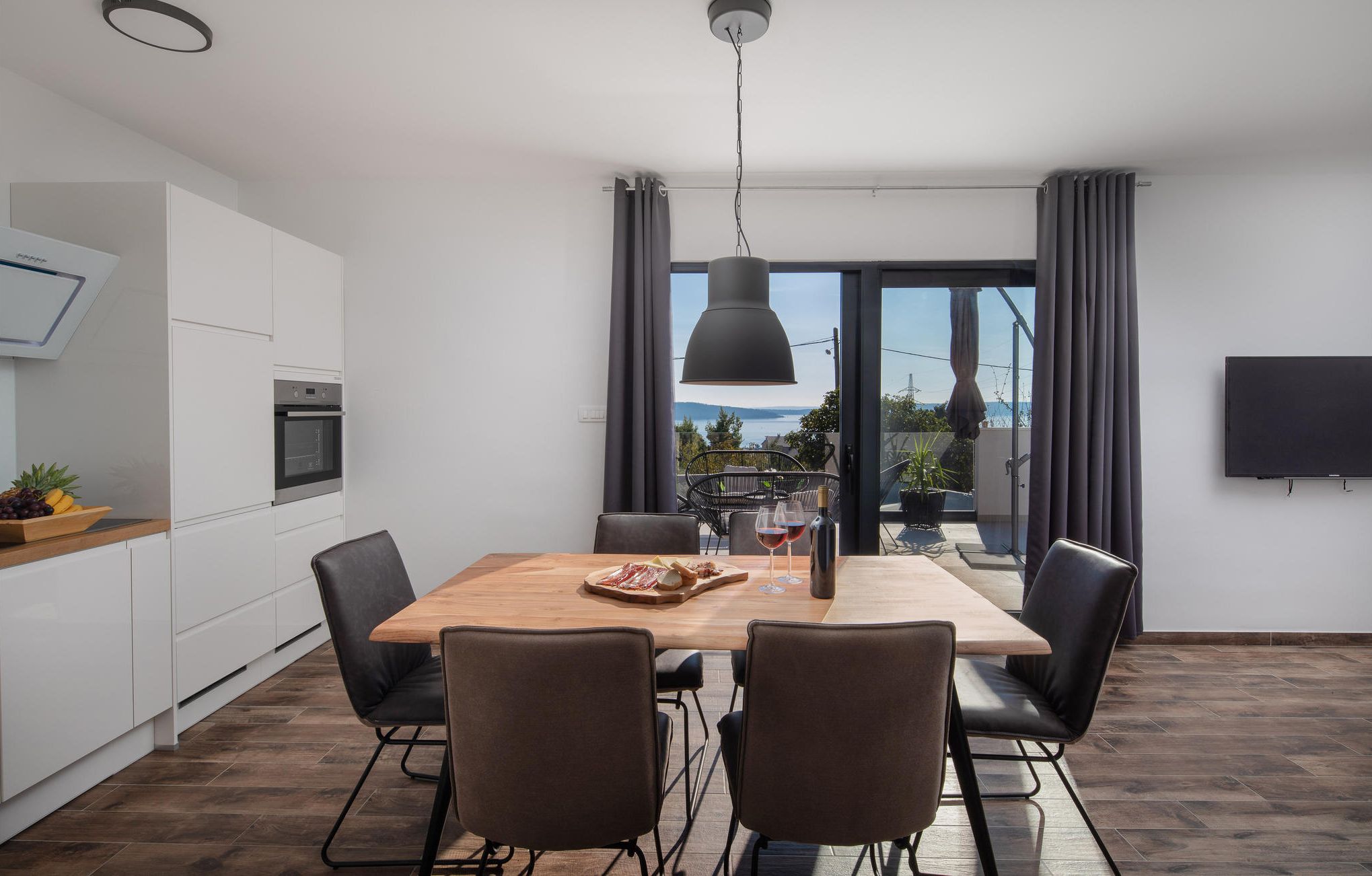 Cozinha moderna com armários brancos, forno embutido e área de jantar com mesa de madeira e cadeiras pretas.