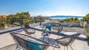 Eine Dachterrasse mit drei Korbstühlen, einem kleinen runden Tisch und Blick auf das Meer und entfernte Gebäude.