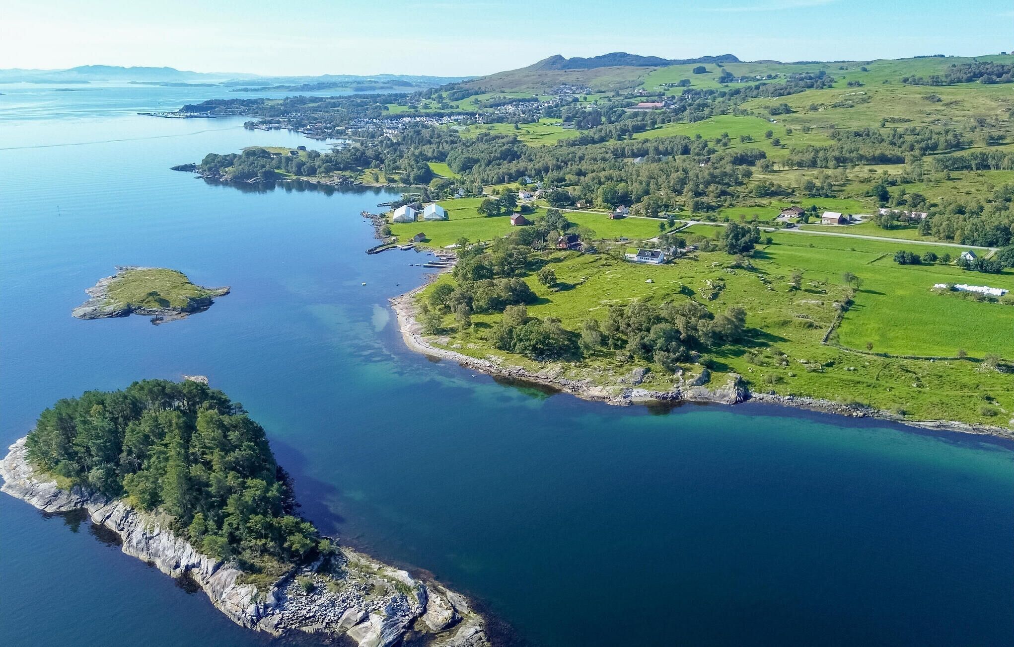 Luftaufnahme einer Küstenlandschaft mit einer kleinen Insel, einem Festland mit verstreuten Häusern und einem klaren, blauen Meer.