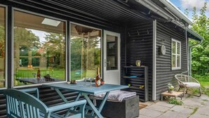 Un espace extérieur accueillant, aménagé avec une table et des chaises, une petite structure en bois et offrant une vue sur un jardin grâce à de grandes portes vitrées.