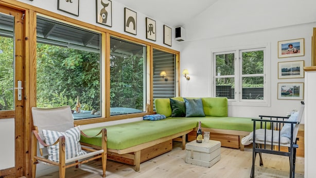 Une pièce chaleureuse avec un banc en bois, un canapé vert et un fauteuil. De grandes fenêtres sont équipées de stores, le sol est en bois et des tableaux encadrés ornent le mur.