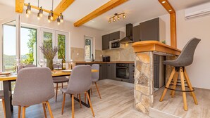 A modern kitchen with a stone accent wall, wooden beams, and a dining area with grey chairs and a wooden table. - 3 bedroom cozy home in Skradin (Skradin)