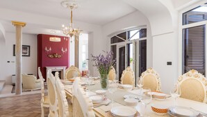 Un comedor con una larga mesa dispuesta para una comida, sillas elegantes, una araña y una chimenea al fondo.