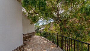 A stone-paved balcony with a black metal railing, overlooking a hillside with trees and a distant view of a building. - Nice home in Antequera (Antequera)