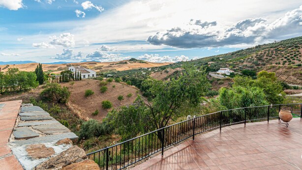 A terrace with stone flooring and a metal railing, overlooking a hilly landscape with scattered buildings and greenery. - Nice home in Antequera (Antequera)