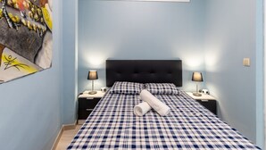 Ein Schlafzimmer mit kariertem Bettbezug, einem schwarzen Kopfteil, zwei Nachttischlampen und einem an der Wand befestigten Kunstwerk.