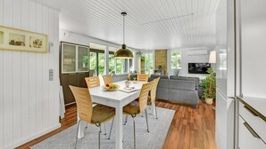 A modern kitchen with white cabinetry, a dining area with a white table and wooden chairs, a grey sofa, and a TV mounted on the wall. - 3 bedroom gorgeous home in Oksbøl (Oksbøl)
