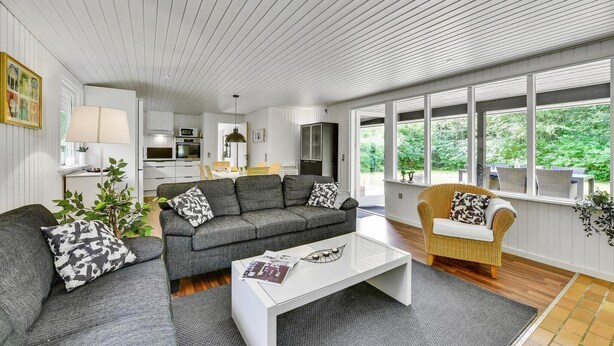 A modern living room with a grey sofa, a white coffee table, and a wicker chair. There is a kitchen in the background with white cabinets and a dining area. - 3 bedroom gorgeous home in Oksbøl (Oksbøl)