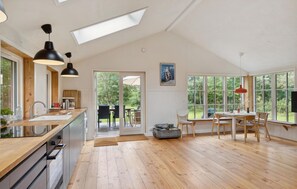 Enjoy a bright and airy space with a high ceiling, kitchen, dining area, and large windows offering views of the surrounding greenery.