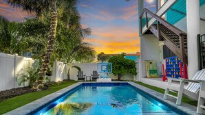 A swimming pool with a view of the sunset, surrounded by a white fence, palm trees, and a house with a blue exterior. - Private POOL/SPA-Almost beachfront, Amazing Gulf Views, walk to Bridge St. (Bradenton Beach)