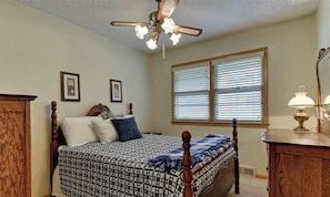 A bedroom with a bed, a ceiling fan, a window with blinds, and a bedside table with a lamp.