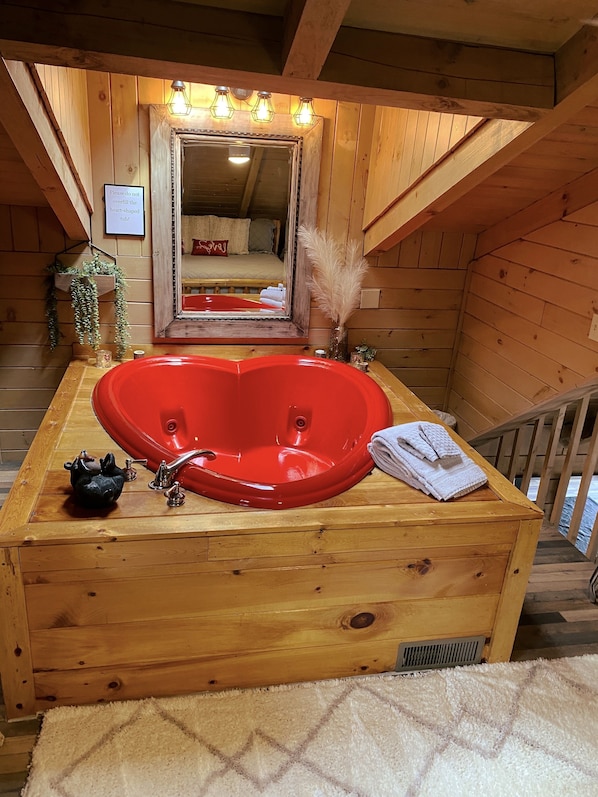 A wooden cabin bathroom with a heart-shaped red bathtub, a small sink, and a mirror above the tub.