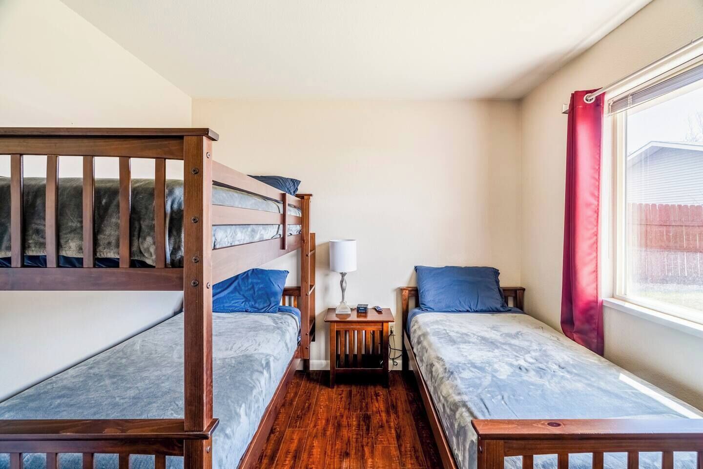 A compact room with two bunk beds and a single bed, a small bedside table with a lamp, and a window with a view of a fence.