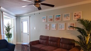 Ein Wohnzimmer mit braunem Ledersofa, einem blauen Sessel und einem kleinen runden Tisch. An der Wand befinden sich gerahmte Bilder und ein Deckenventilator.
