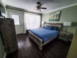 A bedroom with a wooden bed, a ceiling fan, a nightstand with a lamp, and a painting on the wall. - Family friendly condo in Port Aransas (Port Aransas)