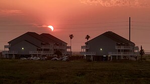 Eine Reihe zweistöckiger Gebäude mit flachen Dächern und Balkonen, gelegen in einer grünen Umgebung, mit einem Sonnenuntergang im Hintergrund.