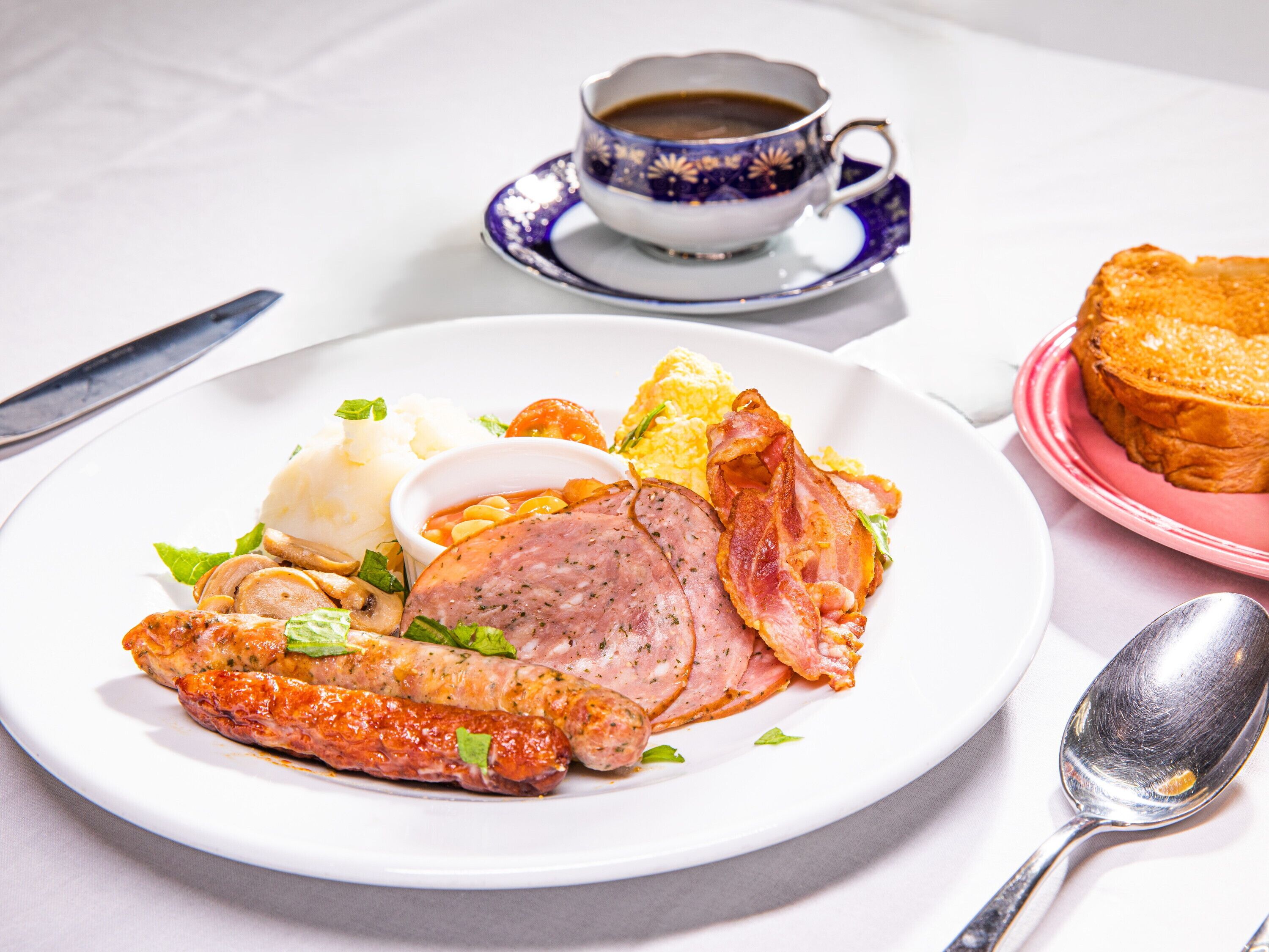 Un plato de comida con salchichas, jamón y tostadas, una taza de café y una cuchara sobre una mesa.