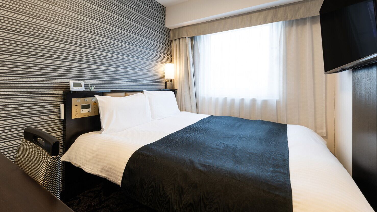 A hotel room with a bed, a bedside table, a lamp, a TV, and a window with curtains.