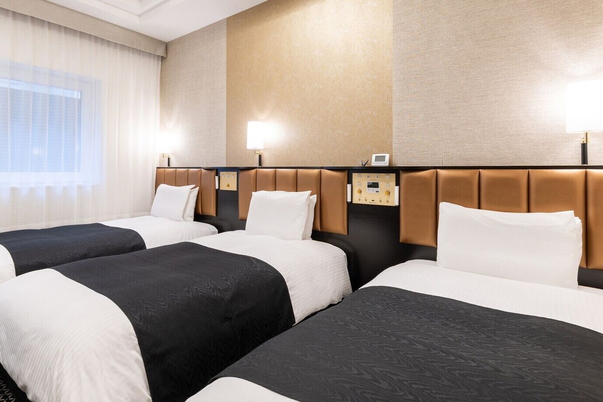 A hotel room with three beds, a control panel, and a window with curtains.