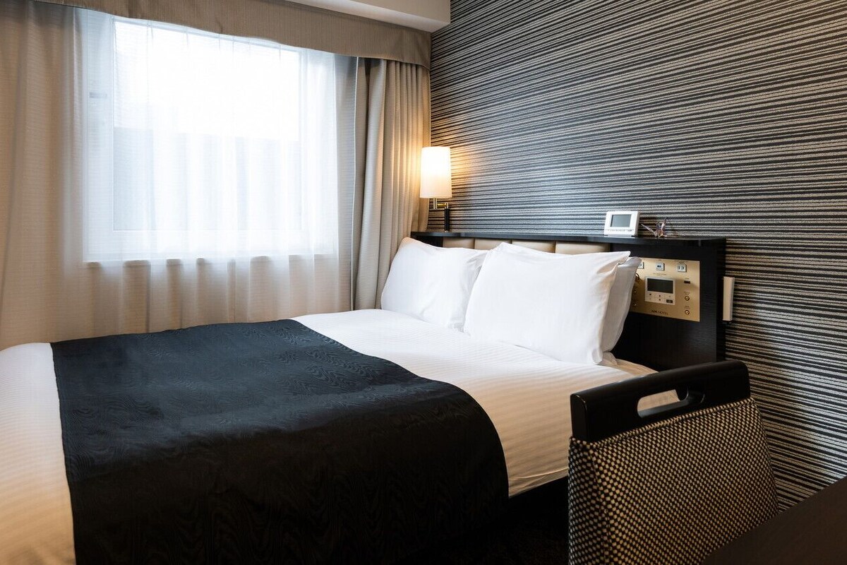 A hotel room with a bed, a TV, a bedside table with a lamp, and a wall-mounted control panel.