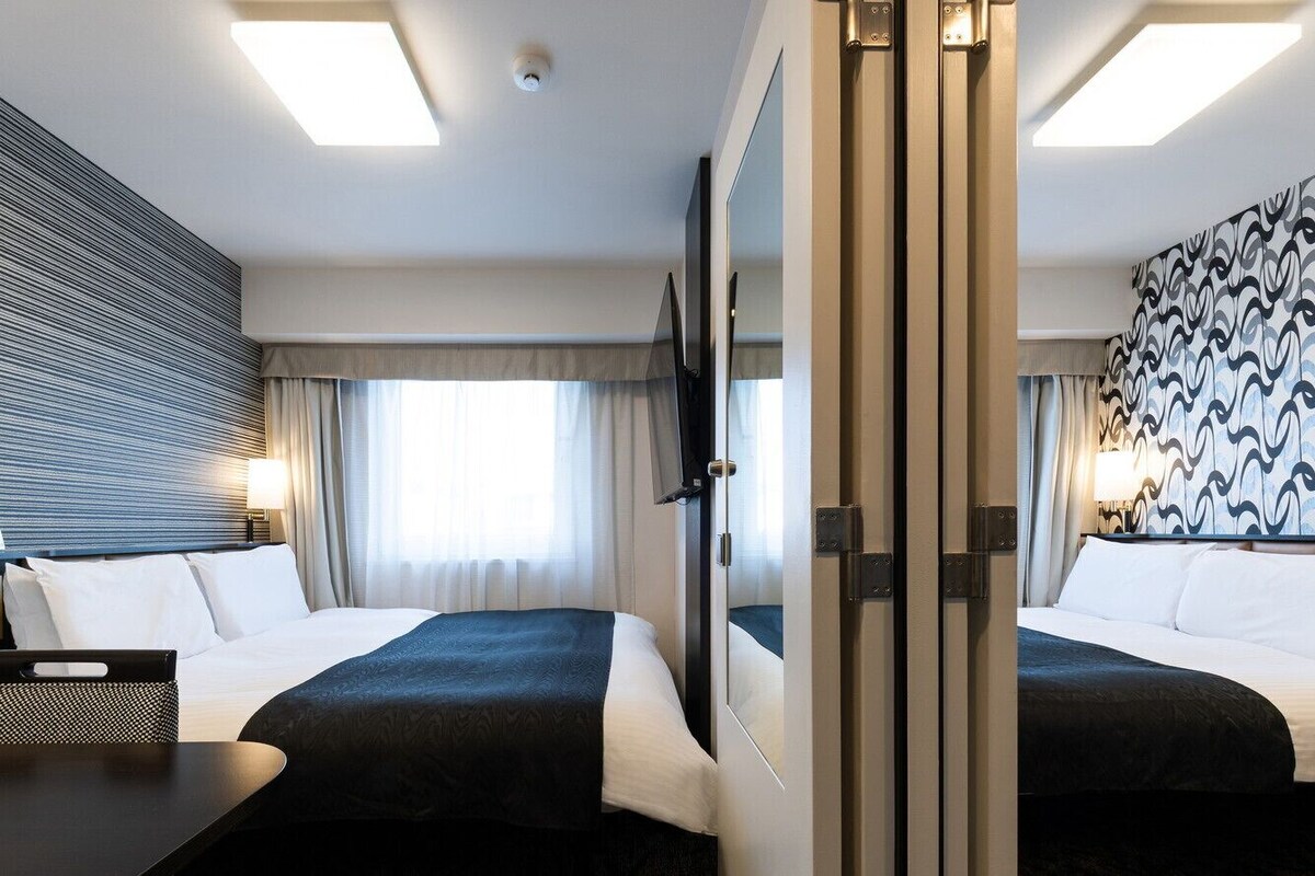A hotel room with two beds, a desk, a control panel, and a patterned wall.
