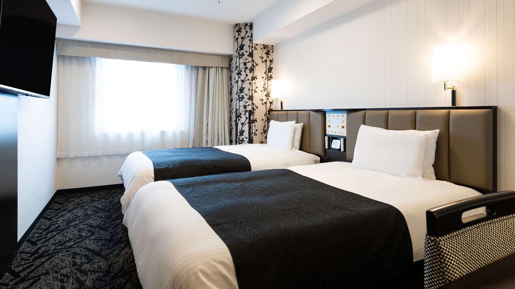 A hotel room with two beds, a TV, a desk with a lamp, and a window with curtains.
