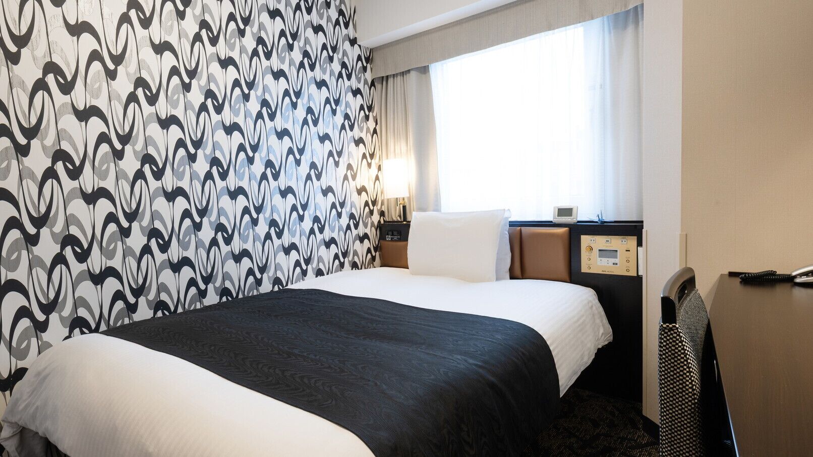 A hotel room with a large bed, a bedside table with a lamp, a control panel, and a window with curtains.