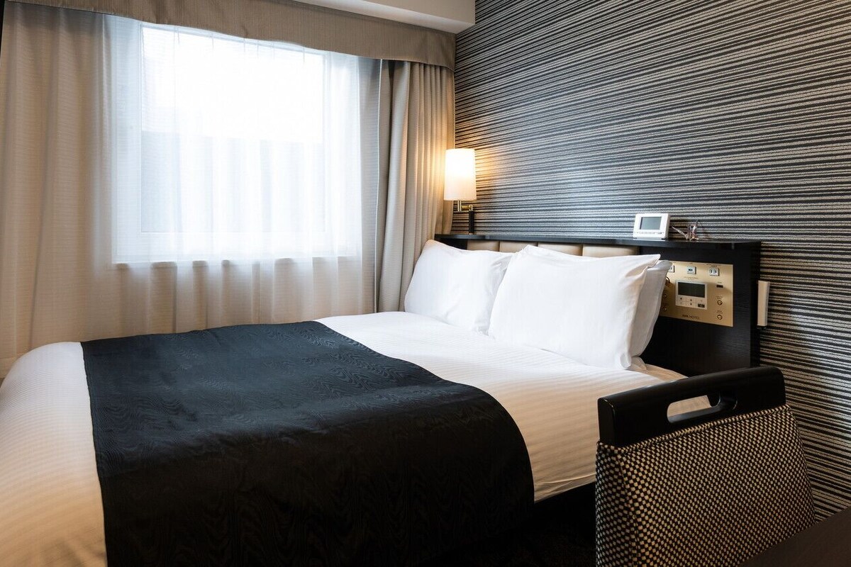 A hotel room with a bed, a TV, a bedside table with a lamp, and a wall-mounted control panel.