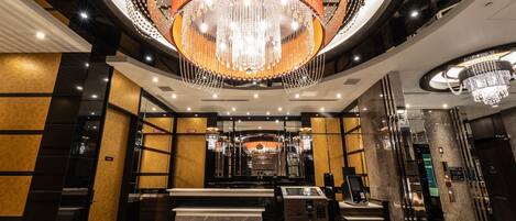 Un hall d’hôtel moderne avec un lustre central, un comptoir d’accueil et un kiosque d’enregistrement.