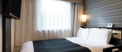 Habitación de hotel con cama, televisión montada en la pared, mesita de noche con lámpara y ventana con cortinas.