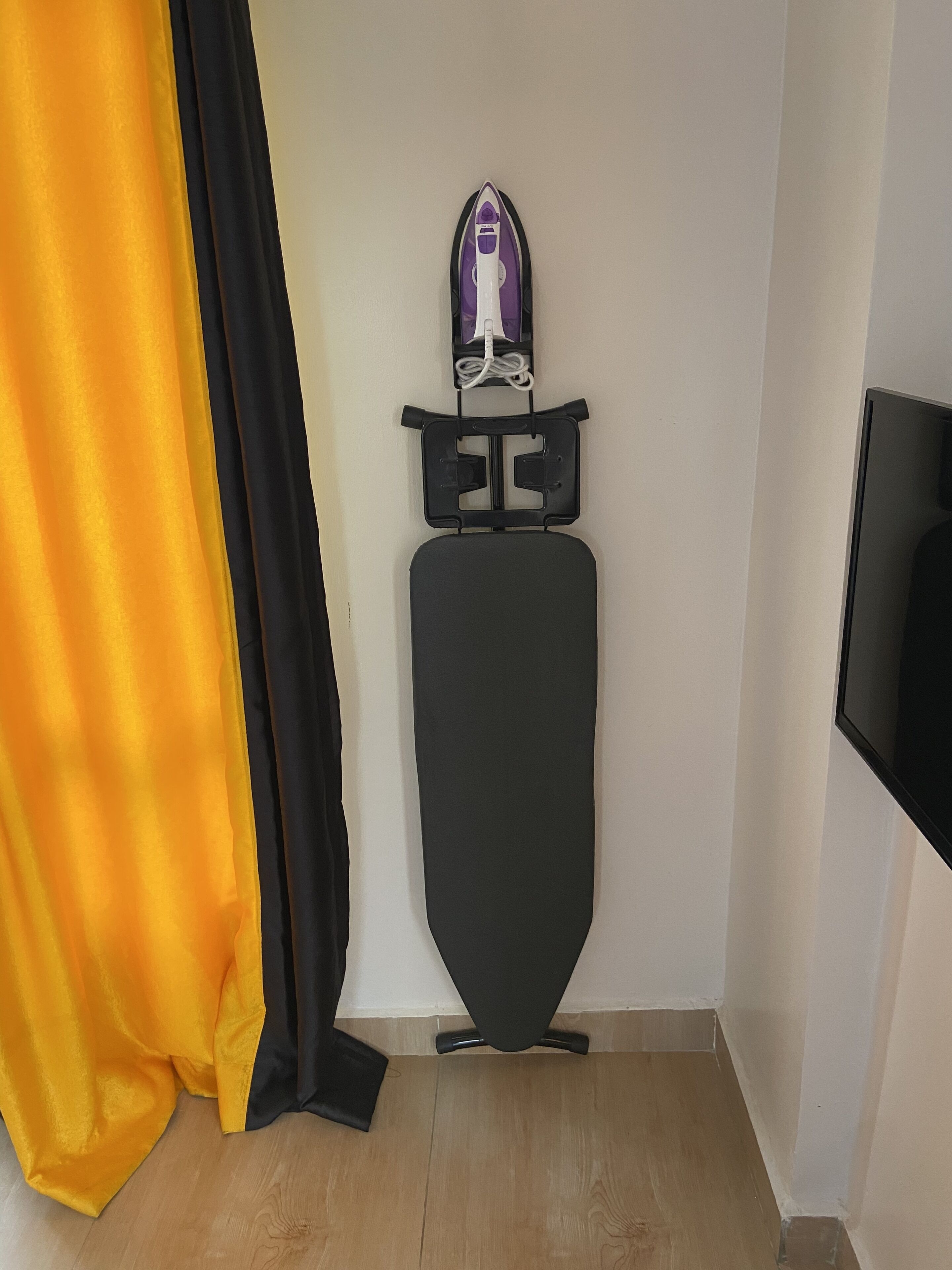 Una habitación con una tabla de planchar negra, un televisor montado en la pared y piso de madera.