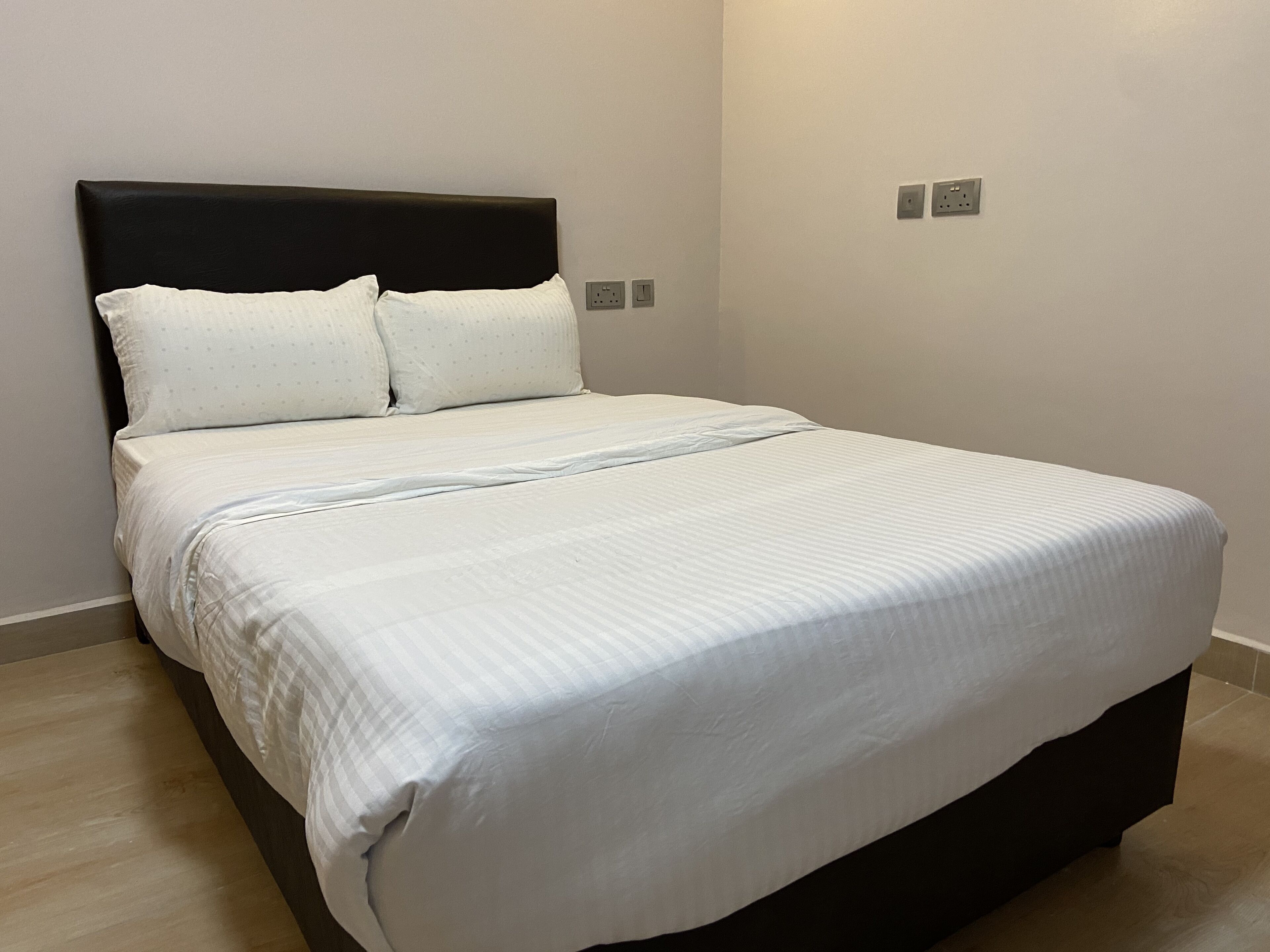 A neatly made bed with white bedding and a black headboard in a room with light-colored walls and a wooden floor.