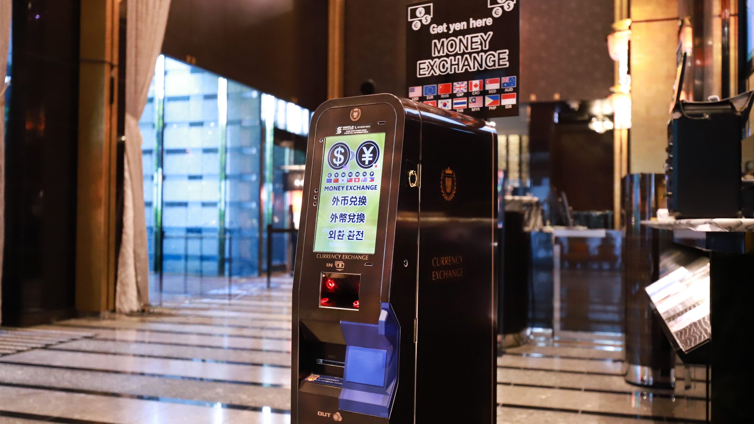 A currency exchange machine with a digital display, located in a well-lit indoor area with a glass facade in the background.