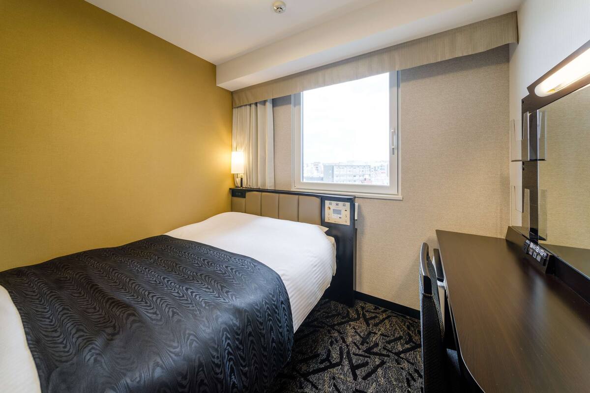 Une chambre d’hôtel avec un lit, un bureau, une chaise, une fenêtre avec des rideaux et un luminaire au plafond.