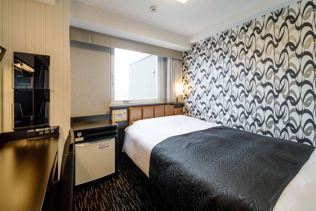 Une chambre d’hôtel avec un lit, un bureau équipé d’un ordinateur, un petit réfrigérateur, une fenêtre et un mur décoré d’un motif.