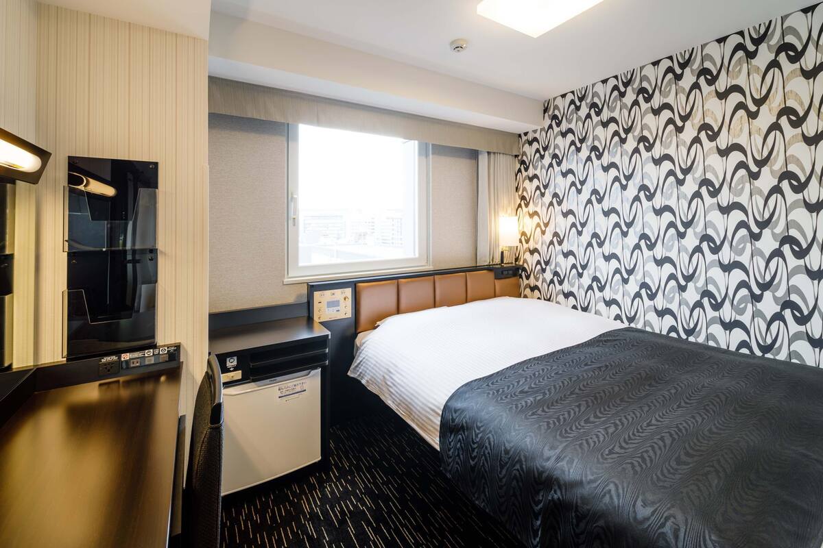 Une chambre d’hôtel comprenant un lit, un bureau avec un ordinateur, un minibar et une fenêtre donnant sur des immeubles.